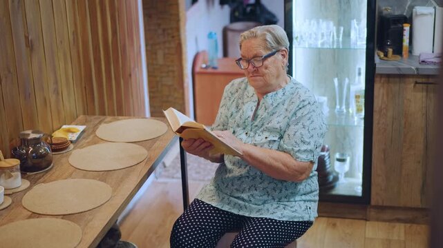 Grandma reading recipe at kitchen table, surrounded by placemats, jars and teacup, preparing breakfast ideas with focused hands, sunlight through window, homestyle atmosphere and vintage tableware,