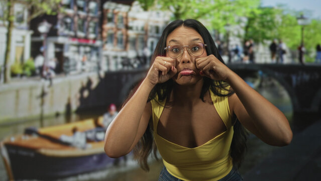 Woman pinching her cheeks with fingers and making a funny face wearing glasses and a yellow tanktop in studio; playful mischief.