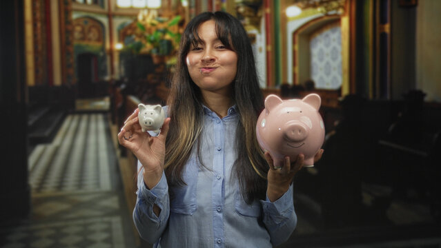 Young brunette latina woman holding a small white and a large pink piggybank with both hands inside a catholic church interior; charity savings.