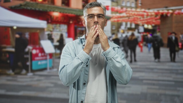 Man with grey hair and glasses blows a kiss hands to mouth on street market lined with red lanterns and stalls; celebration affection.