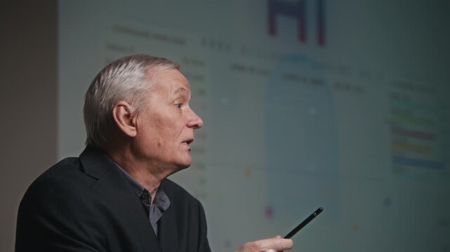 Side view rack focus shot of senior Caucasian male presenter in black suit standing in front of projection screen with graph and giving speech about medical research during conference