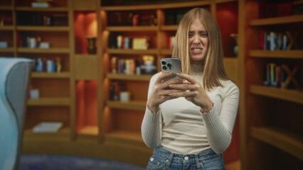 Woman holding smartphone, thumbs typing and frowning in a wooden library with bookshelves and armchair  frustration. © Krakenimages.com