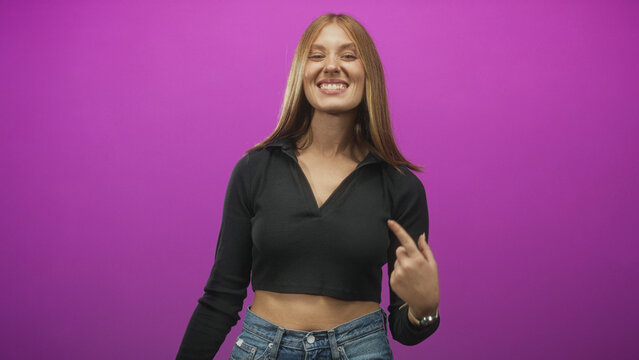 Woman points finger to stomach in purple studio, smiling with exposed midriff and casual jeans; confidence joy playful.
