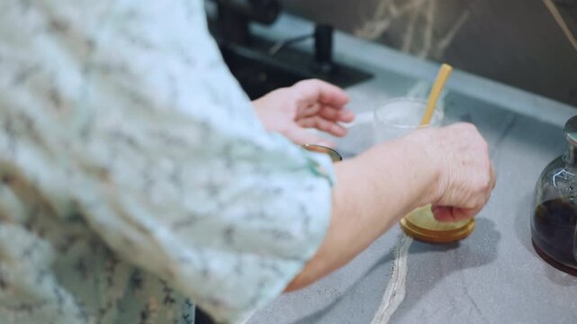 Grandma hands stirring sugar for grandson, closeup of gentle hands using wooden spoon, green glass cup, sugar jar lid on counter, kitchen counter ritual, caregiving mood, morning routine