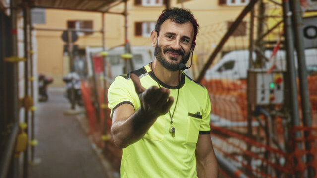 Man referee middle aged bearded with whistle necklace and headset beckons with outstretched hand on a street near scaffolding and safety barriers; friendly greeting.