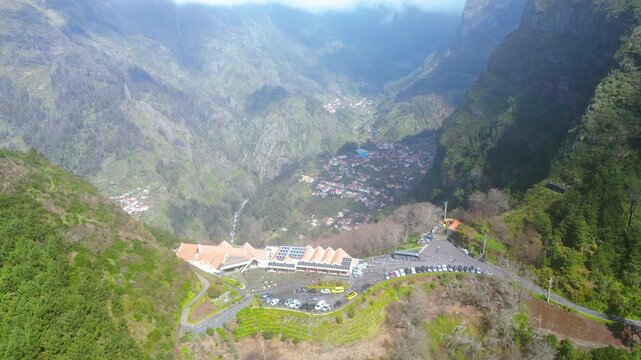Panoramic view of Curral das Freiras, Madeira, nestled in a deep, verdant valley. Terraced slopes and a charming village under midday sun, with a hint of rainbow.
