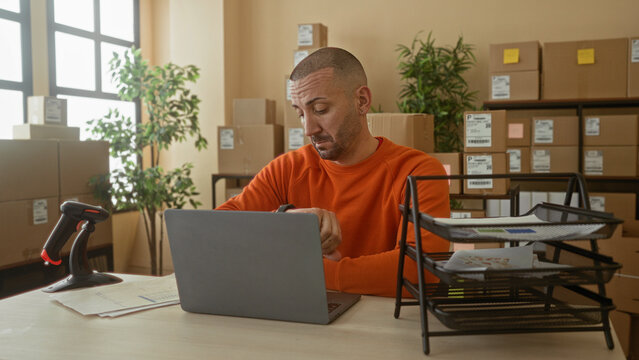 Man in orange sweater checks laptop and watch, hand on chin, barcode scanner on desk in building; small business frustration.