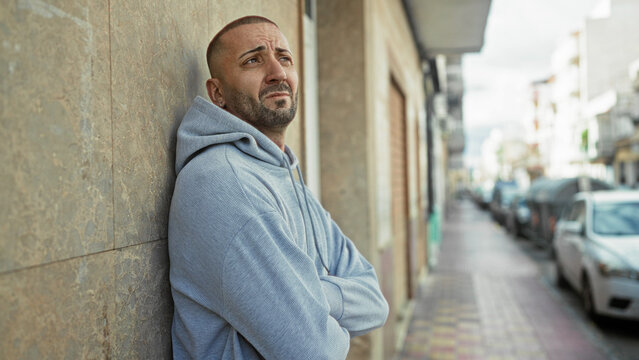 Man leaning against a building with arms crossed, face turned toward the street, wearing a light hoodie and small earring; quiet contemplation.