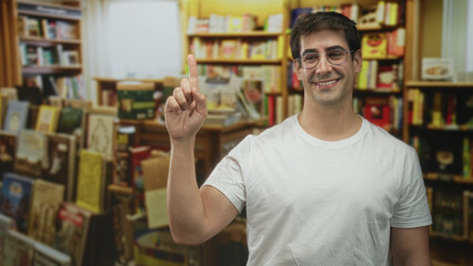 Man in white tshirt and glasses pointing index finger up among crowded bookshelves in a building while smiling  cheerful learning. © Krakenimages.com