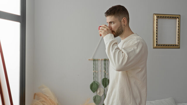 Man holding red mug and sipping coffee beside bed near a window in a bedroom; morning routine serenity.