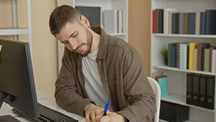 Man writing with blue pen at desktop computer and monitor, leaning forward with hand on keyboard, bookshelves nearby in building  focused productivity. © Krakenimages.com