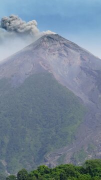 Vertical Screen: Active volcano releases thick gray ash plume into blue sky. Conical peak towers above dense tropical forest covering lower slopes. Volcanic terrain contrasts with lush greenery.