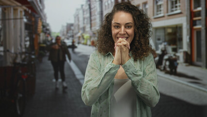 Fototapeta premium Young hispanic woman smiling with fingers crossed and hands pressed together in prayer on a city street lined with shops; hopeful.