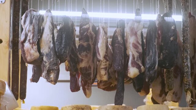 Various cuts of dried, cured meat and sausages hang above wooden shelf lined with several wheels of aged cheese in traditional deli. Dairy storage room on small cheese farm. Food production process