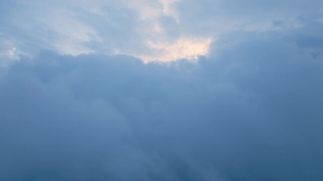 Soft sunlight pierces through thick layers of blue and gray clouds creating a dramatic atmospheric scene. Dense cloud formations drift across overcast sky with subtle warm light emerging from behind.