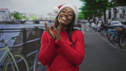 Fototapeta premium Woman wearing santa hat and red sweater smiling and clapping hands on a canal street lined with bicycles and railing; holiday joy warmth.