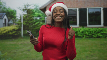 Fototapeta premium Woman in santa hat holding smartphone and giving thumbs up in front of a building, smiling and dancing subtly; holiday cheer joyful.