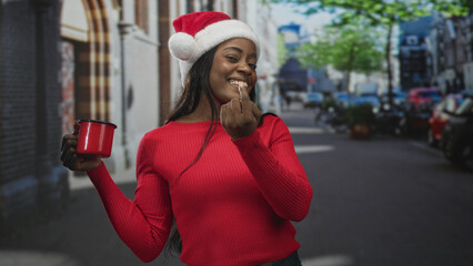 Fototapeta premium Woman in santa hat holding red mug and showing middle finger on a street lined with parked cars and trees, smiling broadly; mischievous.