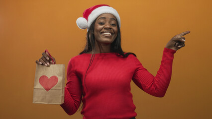 Fototapeta premium Woman in red sweater and santa hat holding a paper gift bag with heart and pointing right while smiling in studio; holiday joy cheer.