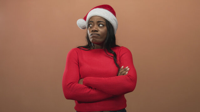 Woman wearing santa hat folds arms in studio with brown backdrop and red sweater visible; holiday annoyance.