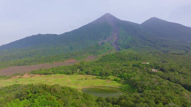 Stunning aerial footage of Guatemala showcasing the active Fuego Volcano rising above dense tropical rainforest. Lava flow path visible on volcanic slopes with misty peaks and a serene pond below.