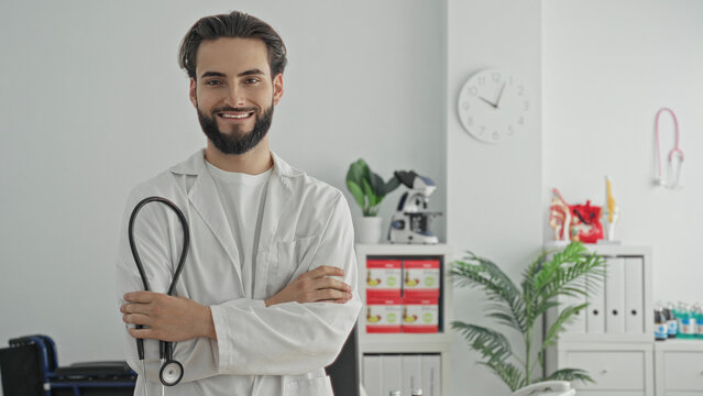 Young hispanic doctor man smiling in white coat, holding a stethoscope with arms crossed in a bright medical clinic consultation room; care confidence.