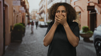 Fototapeta premium Young african american woman covering mouth with both hands on a cobblestone city street, wide eyes and casual black shirt; surprise discovery.