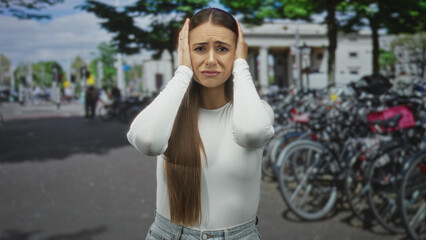 Fototapeta premium Woman covering ears with hands beside crowded bicycle rack on busy city street near columns and trees; urban stress.