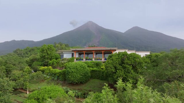 Aerial drone footage reveals an elegant villa with red tile roof surrounded by lush tropical gardens in Guatemala. The active Fuego Volcano emits smoke plumes in the misty mountain background.