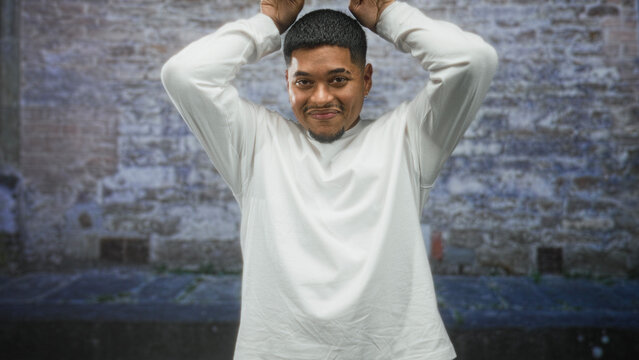 Hispanic man with beard, hands on head gesture and slight smile framed against a weathered brick wall on a street; playful confidence.