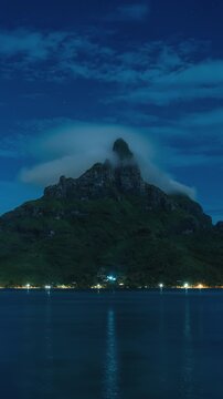 Vertical Screen: Mount Otemanu volcanic peak wrapped in swirling clouds under starry night sky in Bora Bora. Coastal resort lights reflect on calm lagoon waters creating serene tropical atmosphere.