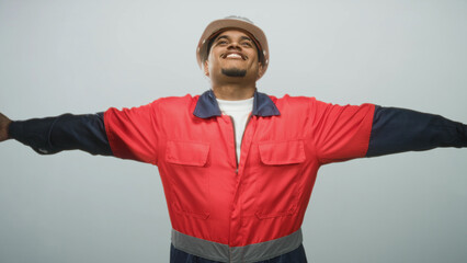 Bearded man in red coveralls and hard hat stands with arms outstretched in studio as construction worker, smiling broadly  pride. © Krakenimages.com