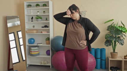 Young plus size woman stands with hand on hip and reaching arm toward exercise ball and mirror, surrounded by yoga mats and potted plant in studio  home workout fatigue. © Krakenimages.com