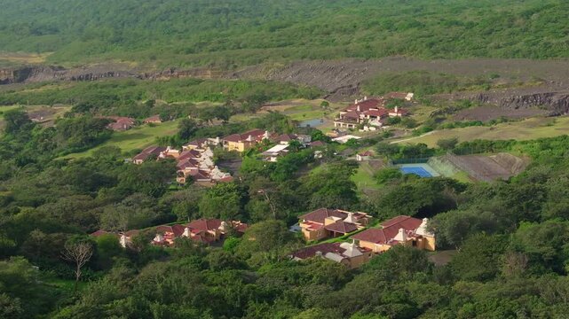 Aerial drone footage captures upscale resort community with terracotta roofed villas nestled among dense tropical forest. Tennis court and sports facilities visible near rocky hillside terrain.