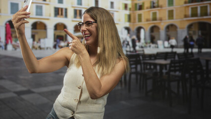 Fototapeta premium Woman holding smartphone points finger to phone at a sunlit building plaza, bare arms visible in sleeveless top while taking selfie; joy travel memories.