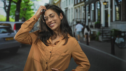 Obraz premium Woman touching hair with hand, smiling while wearing glasses and orange shirt, standing on street with parked cars and row houses; carefree joy.