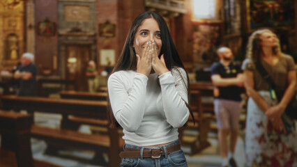 Fototapeta premium Woman covers mouth with hands inside ornate church building by wooden pews and paintings with tourists; awe surprise.