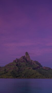 Vertical Screen: Stunning time-lapse of Mount Otemanu in Bora Bora transitioning from starry night to vibrant sunrise. Volcanic peak rises above calm lagoon with resort lights reflecting on water.