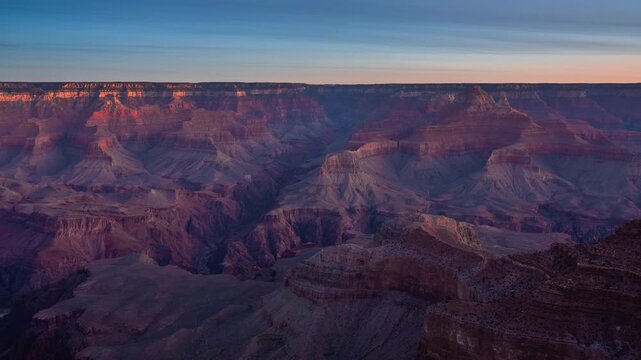 Breathtaking time-lapse captures Grand Canyon at sunset as warm golden light gradually illuminates dramatic layered rock formations while purple and blue shadows deepen across vast Arizona landscape.
