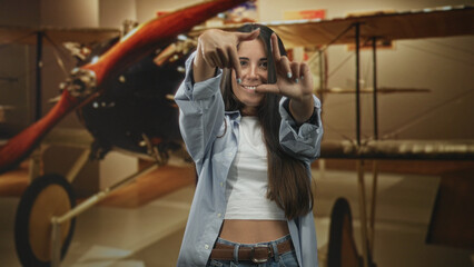 Woman framing a rectangle with her fingers and smiling at an airplane propeller inside a museum building  playful curiosity. © Krakenimages.com