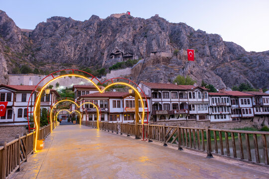 Twilight view of Amasya, Turkey, featuring a glowing arched bridge over Yesilirmak River. Traditional Ottoman houses line the riverbank below the ancient Pontic King Rock Tombs and Amasya Castle on th