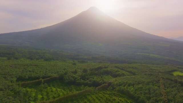 Aerial view of Volcano Arenal in Costa Rica during sunrise with sunlight illuminating the misty peak. Dense tropical rainforest covers the volcanic slopes and surrounding valley in lush green foliage.