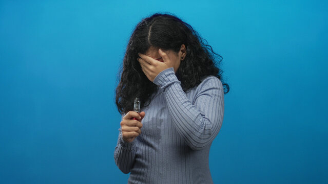 Woman holding a vape device and rubbing her eye in a blue studio while grimacing, visible hand grip on vaporizer near chest level; irritation.