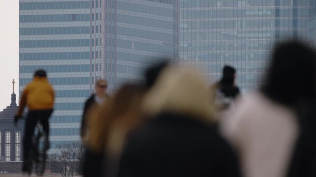 Telephoto view glass skyscraper skyline with strong perspective compression, overcast soft light, glass facade details, blurred silhouettes of people walking in foreground, urban lifestyle slow motion