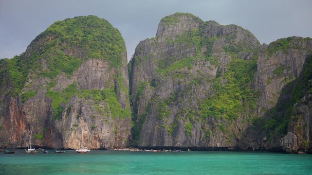 Majestic limestone cliffs draped in vibrant green foliage tower above turquoise tropical waters as traditional longtail boats and sailboats float peacefully near the shore under overcast skies.