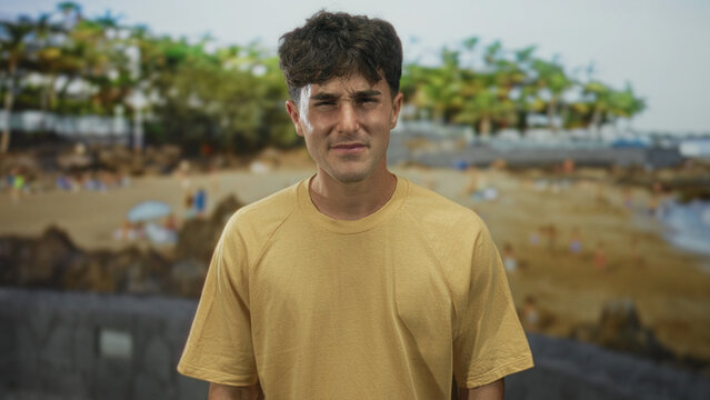 Man in yellow t shirt finger to cheek for toothache on crowded sandy beach by palm trees and seawall, squinting in sunlight; discomfort.