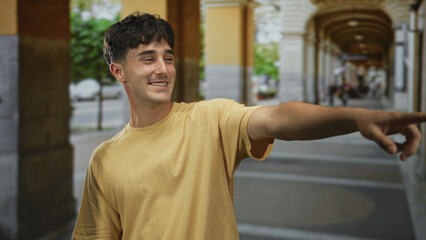 Fototapeta premium Man in yellow t shirt pointing finger with outstretched arm on a street lined with stone columns, looking to his right; playfulness curiosity.