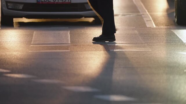 Cinematic close up of urban zebra crossing at dusk, pedestrians walking in slow motion, cars waiting at traffic light creating strong backlight silhouettes on asphalt street