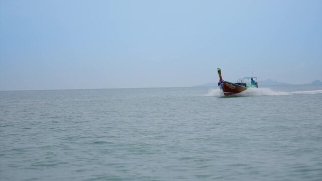 Traditional Thai longtail boat with colorful decorative bow ribbons speeds across calm turquoise ocean waters, creating white spray wake under hazy blue sky with distant mountain islands on horizon.