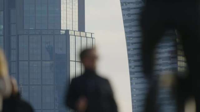 Urban lifestyle scene with blurred foreground pedestrians in motion, sidewalk activity and commuters on their way, distant blue glass skyscrapers skyline in crisp focus under soft daylight